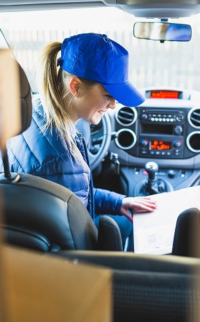 Een lachende vrouw met een blauwe pet en jas zit in een auto, omringd door kartonnen dozen, en bekijkt een document waarin werk als pakketbezorger wordt voorgesteld.