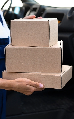 Een persoon in een blauwe overall houdt drie kartonnen dozen vast naast een open bestelwagendeur, wat wijst op pakketbezorging. De scène straalt efficiëntie uit.